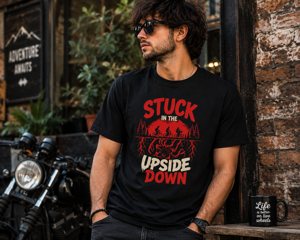 A man wearing a black graphic t-shirt with a red and white design that reads 'STUCK IN THE UPSIDE DOWN' and a red silhouette of a mountain biker, leaning against a brick wall next to a motorcycle.